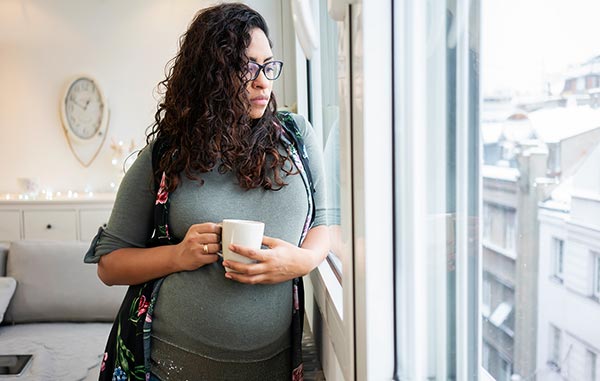 Pregnant woman looks out her window while holding a mug of tea