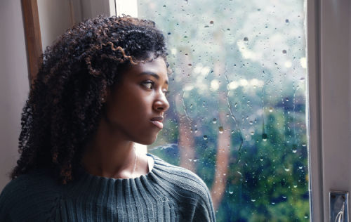 Young woman looks wistfully out her window at the rain
